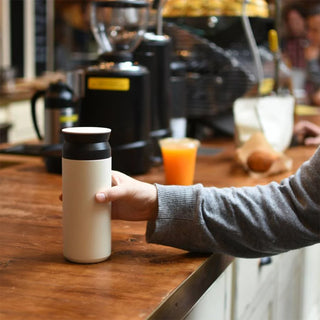 Person holding a white and black insulated mug on a wooden counter with coffee equipment in the background.