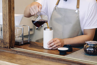 Person pouring coffee from a glass carafe into a white thermos on a wooden counter.