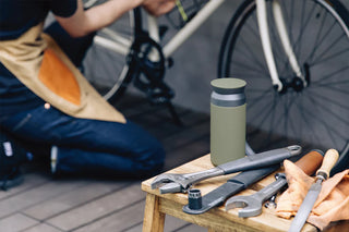 Green water bottle on a wooden table with tools and a person working on a bike in the background.