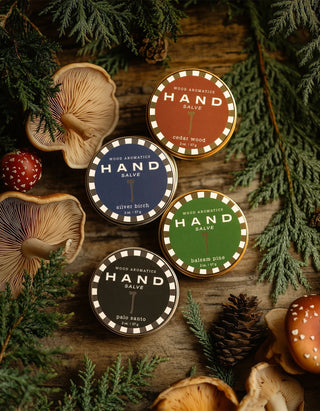Four tins of Wood Aromatics Hand Salve on a wooden surface with greenery and mushrooms.