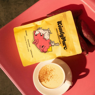 Yellow packet of sweet potato latte mix with cartoon cow design on a red surface next to a cup of coffee.