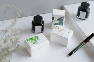 A styled flat-lay featuring two ink bottles, next to white boxes of packaging with various colors, and a dark green fountain pen arranged on a white surface with dried flowers.