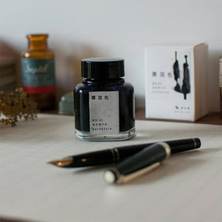 A bottle of dark ink with a black cap and a white label sits on a tabletop, a black fountain pen placed in front