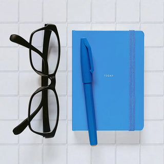 A pair of black eyeglasses and a bright blue pen resting on a blue hardcover notebook with the word 'TODAY' printed on the cover, arranged on a white surface.