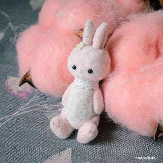 Small plush rabbit toy with a bow tie on a textured surface with pink fluffy objects.