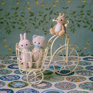 Toy bicycle with a basket containing two plush toys and a teddy bear on top against a decorative background.