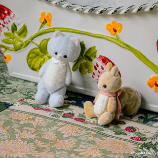 Two plush toys, a cat and a squirrel, sitting on a floral-patterned surface with decorative elements in the background.