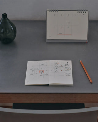 opened notebook calendar with colorful writing with an orange colored pencil on the table. in the background is a desk calendar and vase