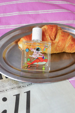Perfume bottle with illustrated colorful design on a metal tray with a croissant, on a striped pink and white background.