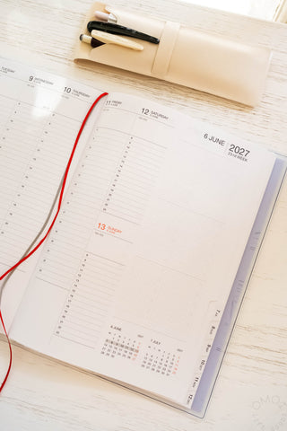 Opened planner page showing weekly vertical view for a date in June 2027 with ribbon bookmarks and a pencil case with items in the background