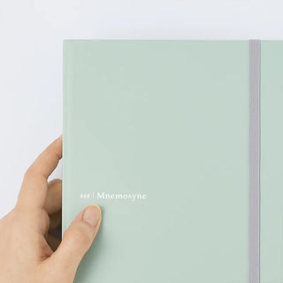 A hand holding a mint green notebook with a white elastic closure band and the text '888 | Mnemosyne' printed on the cover