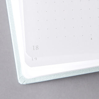 Close-up of a notebook page corner with a pale mint blue textured cover, dotted grid area, and printed page numbers 18 and 14 on a gray background.
