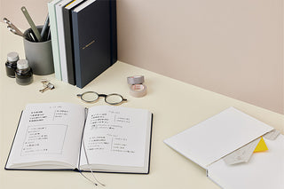 A styled desk with an open notebook showing handwritten notes, other stacked notebooks, eyeglasses, washi tape, small ink bottles, and a pen cup against a warm beige background.