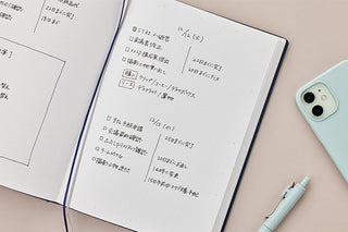 An open notebook with handwritten Japanese notes on white pages, styled on a light pink surface with a mint phone and a matching mint pen nearby.