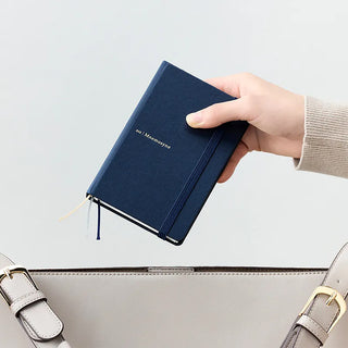 A hand holding a navy blue hardcover notebook with an elastic band, positioned above the top of a light gray bag.