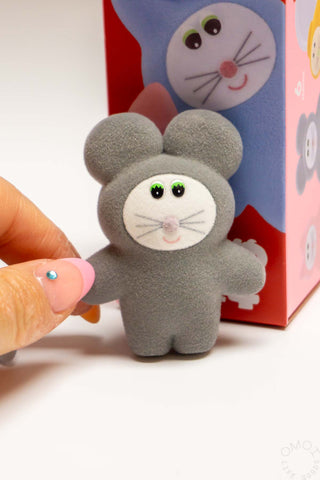 Small gray plush toy held by a hand with a colorful box featuring cartoon characters in the background.