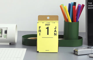 A yellow desk calendar displaying January 1, 2027, on a gray tabletop with office accessories including a green pen cup and other stationery items.