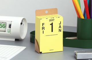 A yellow desk calendar standing upright on a gray tabletop, showing the date 2027-01-01. The calendar has brown cardboard hanger tabs and is surrounded by office items including a white digital clock, a green pencil cup