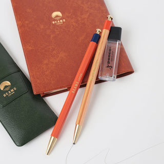 Two mechanical pencils with gold accents on a white surface, next to a red notebook and green pen case with 'Beams' branding.