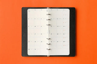 An open 7-day binder calendar with white pages and a black textured cover, held together by silver metal rings, set against a solid orange background.