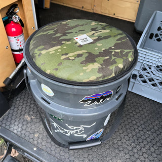 Gray bucket with a camouflage seat pad on a textured surface, surrounded by a fire extinguisher and crates.