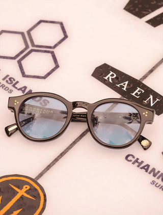 RAEN round sunglasses close-up, black frame with blue lenses and gold stud accents