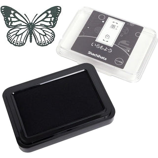 Black Shachihata ink pad shown open with its clear packaging in the background. A butterfly stamp design appears in the upper left 