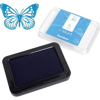 A Shachihata ink pad with blue ink, shown next to its clear plastic packaging with a blue label. A blue butterfly stamp is in the top-left corner