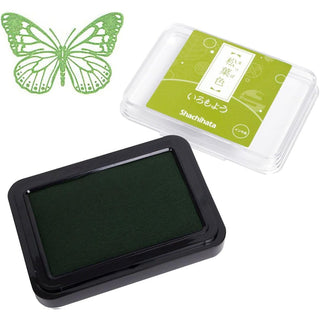 A Shachihata ink pad in dark green shown next to its clear mncontainer with a green label. A green butterfly-shaped stamp in the upper left corner