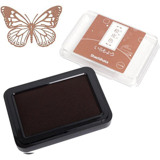 A Shachihata brand ink pad shown open with dark brown ink, alongside its clear plastic case. Next to it is a butterfly-shaped stamp in the upper left