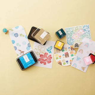 arrangement of colorful stamping tools and printed cards on a beige background, including ink pads, stamps, and patterned cardstock with floral and whimsical illustrations.