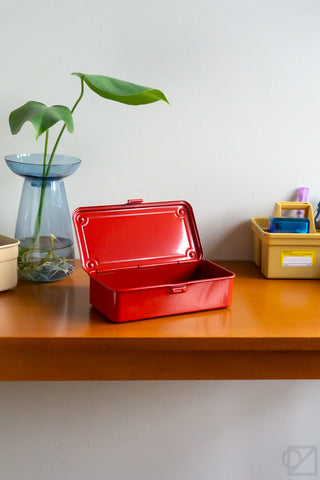 Red metal tool box on a wooden surface with a plant and container in the background