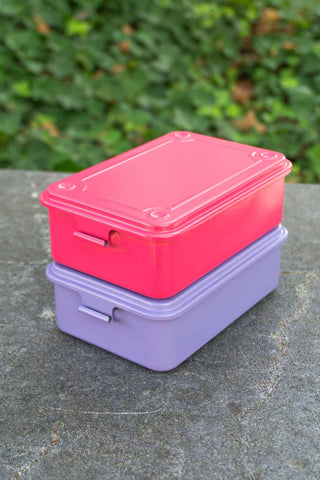 Two stacked metal toolboxes, one pink and one purple, on a stone surface with a green leafy background.