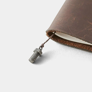 Close-up of a brown leather notebook with a metal screw charm on a light gray background