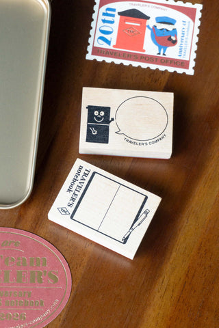 Traveler's Company stationery items including a stamp, card, and notebook on a wooden surface.