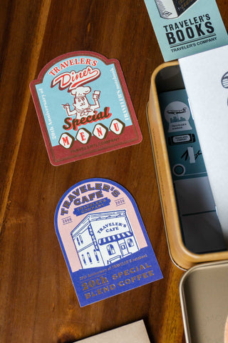 Two 'Traveler's Company' stickers on a wooden surface with packaging in the background