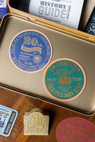 Traveler's company accessories including a metallic tin, stickers, and a clip on a wooden surface.
