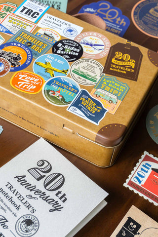 Traveler's Company tin trunk with stickers next to notebook on a wooden surface