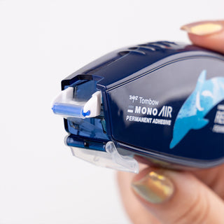 Tombow Mono Air permanent adhesive with a hand holding it on a white background