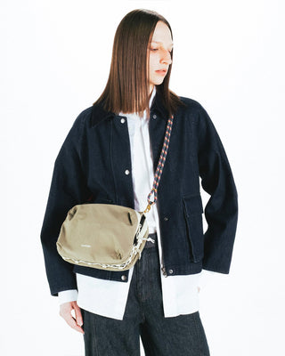 A person wearing a dark indigo denim jacket over a white shirt and dark jeans, carrying a beige canvas crossbody bag with a patterned strap.