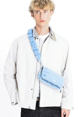 A model wearing a light blue crossbody bag with a matching padded strap, on a white background.