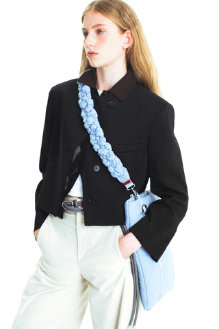 A model wearing a black cropped jacket with white pants, carrying a light blue padded shoulder strap bag, on a solid white background.