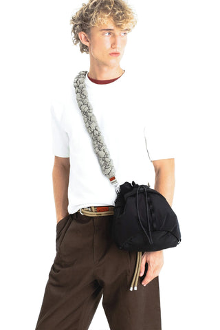 A model wearing a white t-shirt, brown trousers, and a black drawstring crossbody bag with a thick gray padded strap, on a white background.