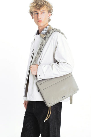 A model wearing a light gray jacket and dark jeans, carrying a light gray puffy messenger bag with a thick, padded light gray shoulder strap.