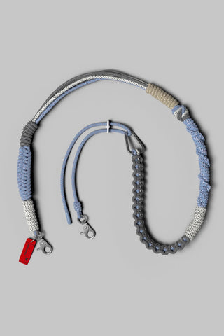 A woven and braided leather strap in shades of gray, light blue, and beige, arranged on a neutral gray background.