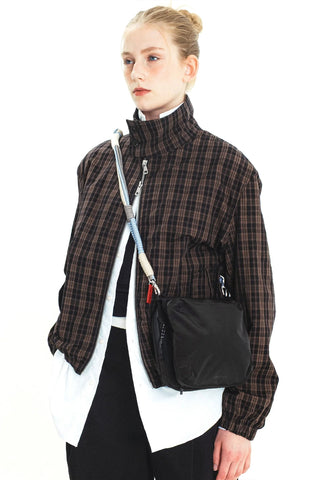 A model wearing a brown and black plaid zip-up jacket carrying a black crossbody bag with a light blue, grey, and beige braided rope strap.