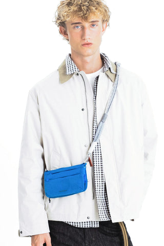 A model wearing a beige jacket carrying a bright blue crossbody pouch bag with a light blue, grey, and beige braided rope strap.