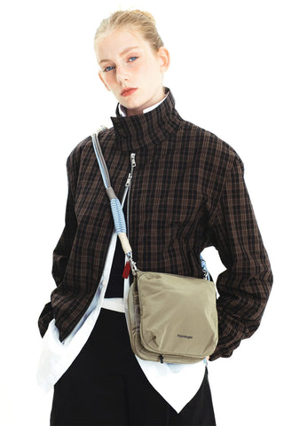 A model wearing a brown and black plaid zip-up jacket, carrying a beige crossbody bag with a blue, grey, and beige braided rope strap over her shoulder, against a white background.