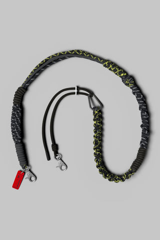 A black and neon yellow braided and knotted rope strap with silver hardware on a neutral gray background.