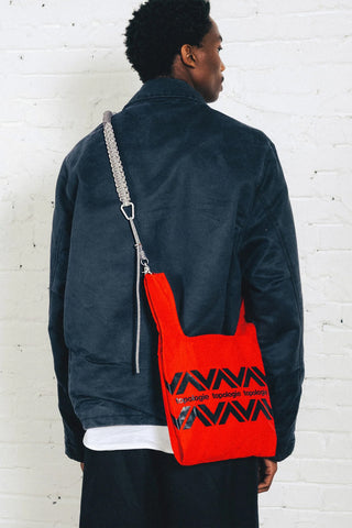 A person shown from the back wearing a navy jacket and black pants, carrying a bright red bag with a grey braided rope strap.